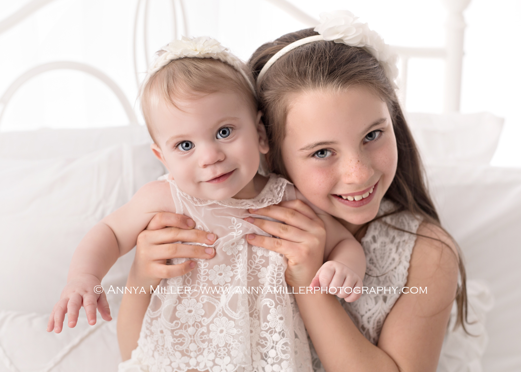 Photograph of baby girl and her big sister on a white bed by Pickering photographer Annya Miller
